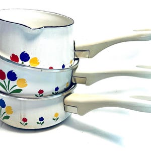 May include: A stack of three white enameled metal saucepans with a pouring spout on the top pan. Each pan features a repeating floral pattern of red, blue, and yellow tulips with green stems and leaves, paired with off-white plastic handles.
