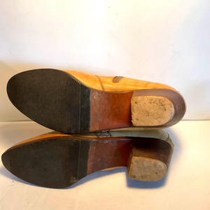 May include: A pair of light tan leather boots with a stacked heel. The soles are dark brown rubber with a textured pattern. The boots have a side zipper and a pointed toe. The heels are approximately 2 inches high.
