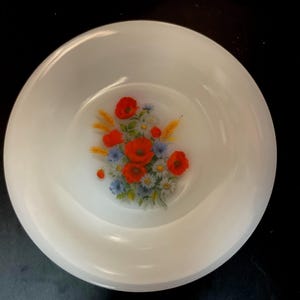 May include: White, round bowl with a floral design in the center. The design features red poppies, blue flowers, and yellow wheat stalks. The bowl is set against a dark background.