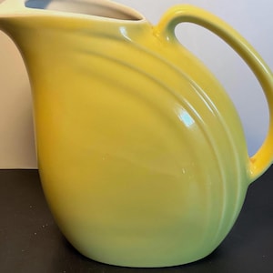May include: A bright yellow ceramic pitcher featuring a smooth, glossy finish, a curved handle, and a wide pouring spout. The body of the pitcher is decorated with three distinct, raised concentric ring details that follow the curve of the handle.