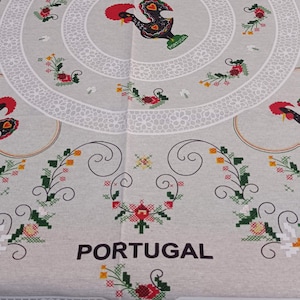 Może przedstawiać: Biały obrus z czerwonym kogutkiem i haftem kwiatowym. Obrus ma wyszyte czarne słowo "PORTUGAL".