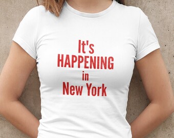 Puede incluir: Camiseta blanca con texto rojo que dice "It's Happening in New York".