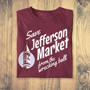 May include: A maroon t-shirt with a white graphic design that says "Save Jefferson Market from the wrecking ball". The graphic also includes a white silhouette of a building with a clock tower.