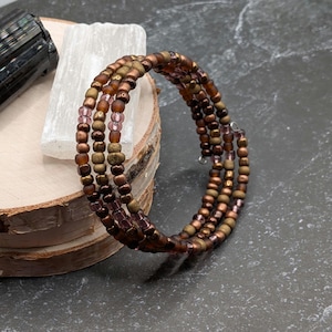 May include: A multi-strand wrap bracelet constructed from small, round beads in alternating shades of metallic bronze, matte brown, and translucent light purple, arranged in a repeating pattern along a flexible wire coil structure.