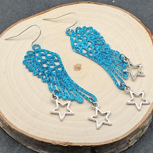 May include: A pair of bright blue glittery wing-shaped earrings featuring a delicate openwork pattern. Each earring is suspended from a silver-toned hook and adorned with two small, dangling silver-toned star charms at the bottom edge.