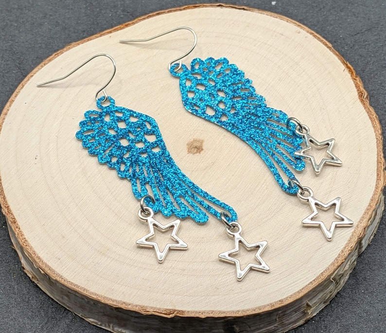 May include: A pair of bright blue glittery wing-shaped earrings featuring a delicate openwork pattern. Each earring is suspended from a silver-toned hook and adorned with two small, dangling silver-toned star charms at the bottom edge.