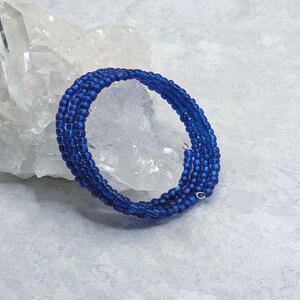 May include: A multi-strand wrap bracelet composed of small, vibrant royal blue glass beads. The bracelet features a flexible, coiled design that circles multiple times, with small silver-toned metal loops visible at the ends of the bead strands.