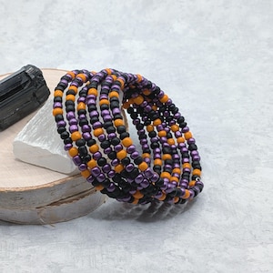 May include: A multi-strand wrap bracelet composed of small, round beads in alternating colors of matte black, vibrant orange, and metallic purple. The bracelet is coiled in a circular shape, showcasing a repeating pattern of these three distinct bead colors throughout the entire length of the accessory.