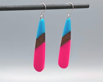 May include: A pair of drop earrings featuring a teardrop shape with three distinct horizontal sections: a bright turquoise top, a dark brown wood grain center, and a vibrant hot pink bottom, suspended from silver-toned metal hooks.