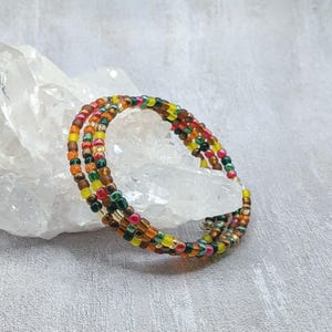 May include: A multi-strand wrap bracelet composed of small glass seed beads in a vibrant palette of yellow, orange, red, green, and brown. The beads feature a mix of opaque, translucent, and metallic finishes, arranged in a repeating pattern.