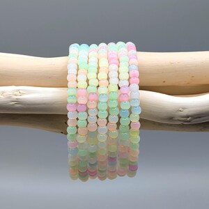 May include: A stack of pastel-colored beaded bracelets. The bracelets are made of small, round beads in shades of pink, yellow, green, and blue. The bracelets are resting on a light-colored wooden branch.