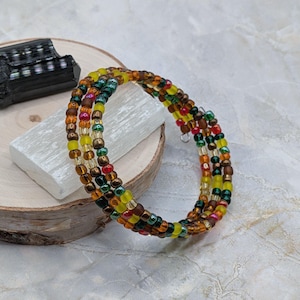 May include: A multi-strand wrap bracelet composed of small glass beads in shades of yellow, orange, brown, red, and dark green. The bracelet features a flexible coiled design and is displayed on a white rectangular stone block.