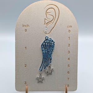 May include: A blue glittery wing-shaped earring featuring two dangling silver-toned star charms. The earring is displayed on a wooden card with printed inch and centimetre scales, showing the wing length is approximately 2 inches or 5 centimetres.