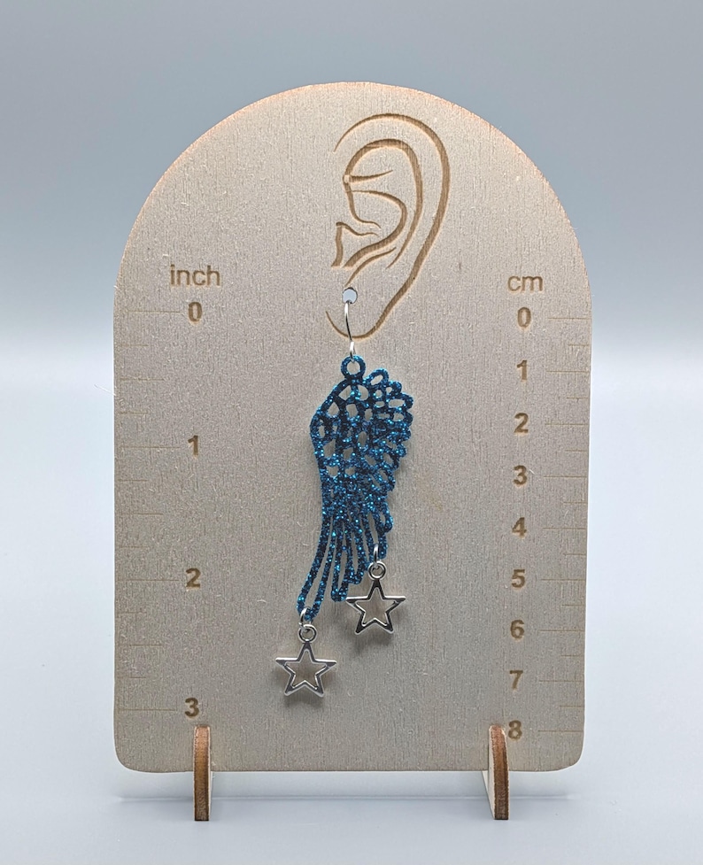 May include: A blue glittery wing-shaped earring featuring two dangling silver-toned star charms. The earring is displayed on a wooden card with printed inch and centimetre scales, showing the wing length is approximately 2 inches or 5 centimetres.