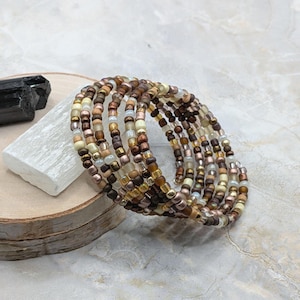 May include: A multi-strand wrap bracelet featuring small glass beads in shades of brown, bronze, cream, and metallic gold. The bracelet is constructed with a coiled design, showcasing a repeating pattern of earthy tones and reflective finishes.