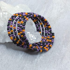 May include: A multi-strand wrap bracelet constructed from small, round beads in alternating colors of matte black, metallic purple, and bright orange. The bracelet features a coiled design that creates multiple layers when worn around the wrist.