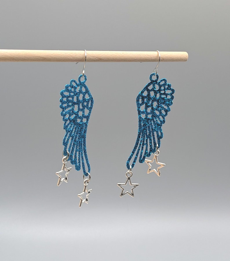 May include: A pair of drop earrings featuring blue glitter-covered wing-shaped cutouts. Each wing is suspended from a silver-toned hook and accented with two small, open-frame silver-toned star charms hanging from the bottom edge of the wings.