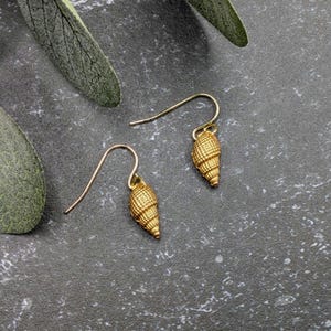 May include: A pair of gold-toned drop earrings featuring a textured, conical seashell-shaped charm suspended from a curved ear wire. The surface of each charm displays a detailed, repeating grid pattern that mimics the natural spiral structure.