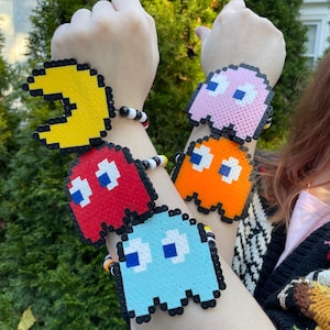 May include: Four colourful pixel art bracelets featuring characters from the classic video game Pac-Man. The bracelets are made of black beads and feature Pac-Man, Blinky, Inky, and Clyde.