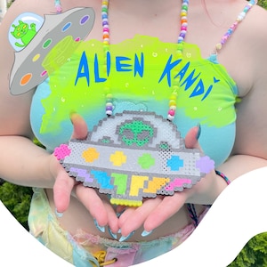 Pode incluir: Uma pessoa vestindo um top azul e verde com o texto "Alien Kandi" nele. Ela está segurando um UFO de pixel art colorido com um alienígena verde dentro.