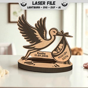 May include: A laser-cut wooden stork figurine, featuring a baby in a pouch. The design includes the name "Emilia", the date 17.07.2025, and the baby's weight, 3327g. The text "10:59 Uhr" is also included. The figurine is two-tone.