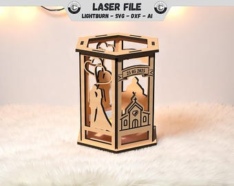May include: Laser-cut wooden lantern with a hexagonal top. The lantern features cut-out designs of a bride and groom, a church, and the date 25 05 2025. The text "LASER FILE" is at the top. The lantern is made of light-colored wood.