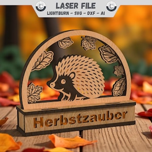 Könnte beinhalten: Eine Holz-Laser-Cut-Dekoration mit einem Igel-Design in einem bogenförmigen Rahmen mit Blattausschnitten. Die Basis lautet "Herbstzauber". Das Design ist vor einem Hintergrund aus Herbstblättern platziert und schafft ein saisonales Thema.