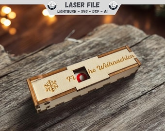 Pode incluir: Uma caixa de madeira com um desenho de floco de neve e o texto "Frohe Weihnachten" na parte superior. A caixa tem um pequeno círculo vermelho recortado na lateral.