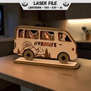 Könnte beinhalten: Ein lasergeschnittenes Wohnmobil-Dekorationsobjekt aus Holz mit einer mehrschichtigen Berg- und Waldlandschaft in den Fenstern, einer Kompassgrafik und dem Schriftzug #VANLIFE. Das Modell steht auf einem Sockel und zeigt helle und dunkle Holztöne.