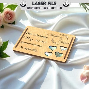 Puede incluir: Una tarjeta de boda de madera con el texto "Der schönste Weg ist der gemeinsame 30. August 2025" y los nombres "Anabel & Rafael". La tarjeta tiene recortes en forma de corazón y está decorada con elementos florales. Rosas y una cuchara de plata en el fondo.