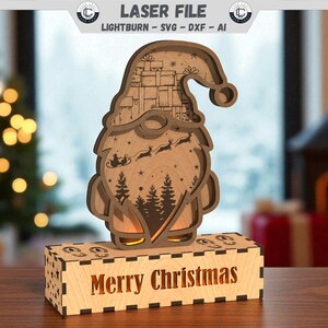Könnte beinhalten: Ein weihnachtliches Wichtel-Licht aus Holz mit der Aufschrift "Merry Christmas" auf dem Sockel. Der Wichtel hat einen Bart, eine Mütze und ein mehrschichtiges Design mit einem Weihnachtsbaum und Rentieren. Das Licht ist für das Laserschneiden konzipiert.