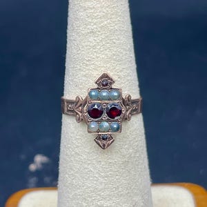 May include: A vintage-style ring with a copper-colored band. The ring features a central rectangular design with two dark red stones and several smaller, light-colored stones. The ring is displayed on a white ring holder.