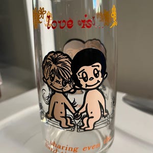 May include: Clear glass with the words "love is!" in red and gold floral accents. The glass features a cartoon illustration of two figures holding hands. The phrase "sharing even hard times" is printed at the bottom.