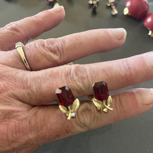 May include: A pair of vintage clip-on earrings featuring rectangular, faceted, deep red gemstones. The gold-toned metal setting includes delicate leaf and small iridescent crystal accents. Other jewelry pieces are visible in the background.