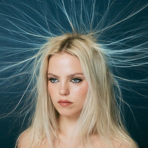 May include: A woman with blonde hair styled in a dramatic, spiky fashion. Her hair is standing on end, creating a halo effect around her head. She is wearing minimal makeup, with defined eyeliner and a neutral lip color. The background is a deep blue.