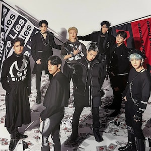 May include: A group of eight people wearing black clothing and posing in front of a white background. They are holding flags with the words "More Lies" and "Make a Move" printed on them. The flags are scattered on the floor around them.