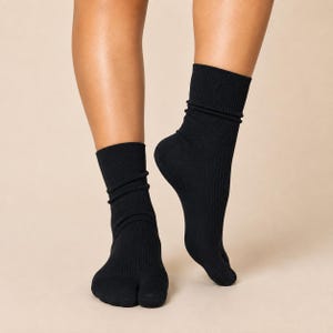 May include: A pair of black ribbed tabi socks featuring a distinct split-toe design that separates the big toe from the other toes. These mid-calf length socks are made of a solid black knit fabric with a vertical ribbed texture throughout the leg.