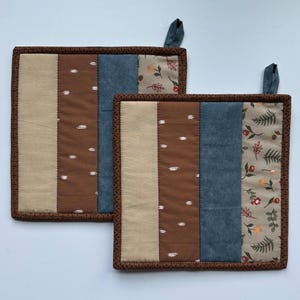 May include: Two brown and beige striped pot holders with blue and floral patterned fabric. The pot holders have a brown stitched border and a loop for hanging.