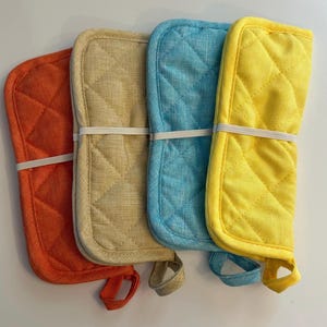 May include: Four quilted oven mitts in orange, tan, turquoise, and yellow. Each mitt is secured with a white elastic band and has a loop for hanging. The mitts have a diamond pattern.