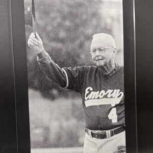 May include: Black and white framed photograph of a man in a baseball uniform. The jersey says "Emory" and has the number 4. The man is holding a baseball cap up in the air.