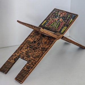 May include: A wooden book stand with a hand-painted religious icon. The stand is made of dark wood and features intricate carvings on the base. The top panel displays a colorful image of an angel with a sword. The stand is designed to hold a book.