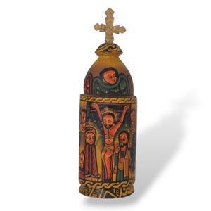 Op de afbeelding: Een handbeschilderd religieus houten artefact met een kruis erop. De cilindrische behuizing toont een afbeelding van een kruisigingsscène met figuren in levendige kleuren. Het bovenste gedeelte heeft een geschilderde engel.