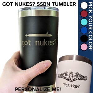 Puede incluir: Un vaso de acero inoxidable negro con una silueta de submarino y el texto "got nukes?" El vaso está siendo sostenido por una mano. El fondo muestra una variedad de opciones de color para el vaso.