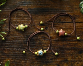 May include: Three star-shaped ceramic charm bracelets with green beads on brown string. The bracelets are on a wooden surface.