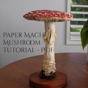 May include: A paper mache mushroom with a red and white spotted cap, a cream-colored stem, and a dark brown base. The mushroom is mounted on a round wooden base. Text on the image reads "PAPER MACHE MUSHROOM TUTORIAL - PDF".