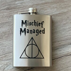 Könnte beinhalten: Eine Edelstahl-Flachmann mit Klappverschluss. Auf der Vorderseite steht der Schriftzug "Mischief Managed" in einer stilisierten Schriftart über einem schwarzen Symbol der Heiligtümer des Todes. Der Flachmann ist rechteckig.