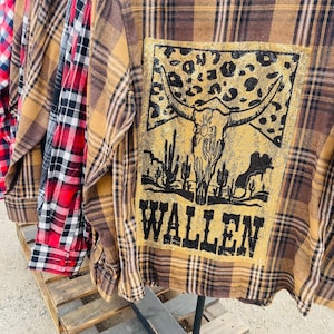 May include: A rack of plaid button-down shirts. The primary shirt is brown and black plaid with a gold graphic on the back featuring a bull skull, desert landscape, and the word "WALLEN". Other shirts are red and black plaid.