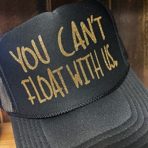 May include: A black trucker hat with a gold glitter design that says "You Can't Float With Us".