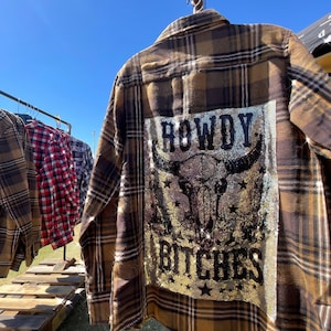 May include: Brown and tan plaid shirt with a sequined design on the back that reads "ROWDY BITCHES" with a bull skull graphic. The shirt is hanging on a metal rack with other plaid shirts in the background.