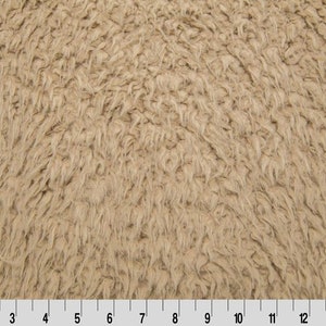 May include: Close-up of a tan, shaggy fabric with a textured, fur-like appearance. The material has a dense, uneven surface with long, soft fibers. A ruler is at the bottom, indicating measurements in inches.